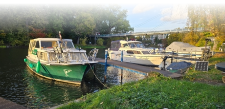 Mehrere Motorboote liegen an den Gastliegeplätzen eines grünen Uferbereichs unterhalb einer Fußgängerbrücke an der Saale.
